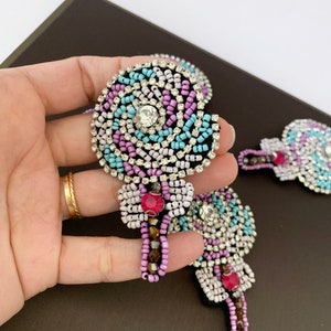 Puede incluir: Un conjunto de tres parches con forma de piruleta, adornados con cuentas y joyas de colores. Los parches están hechos con una variedad de cuentas, incluyendo transparentes, rosas, moradas, azules y marrones. Cada parche presenta un gran strass transparente en el centro de la piruleta.