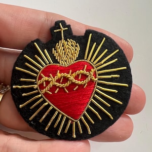 以下が含まれることがあります： 黒いフェルト地に、金糸で刺繍された赤いハート。金色の光輪で囲まれ、茨の冠が飾られています。