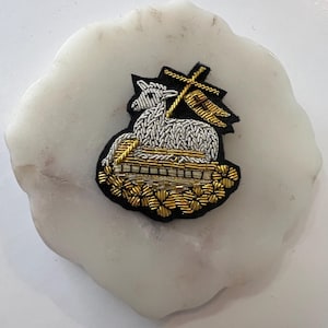 May include: A white lamb with a gold cross and flag embroidered on a black background. The lamb is sitting on a gold and silver embroidered base.