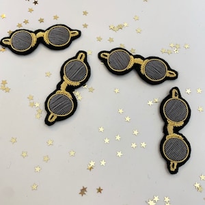 以下が含まれることがあります： 黒い背景に金色のステッチが施された丸いサングラスの刺繍パッチが4つ。サングラスは、金色のキラキラした星が散りばめられた白い背景の上に置かれています。