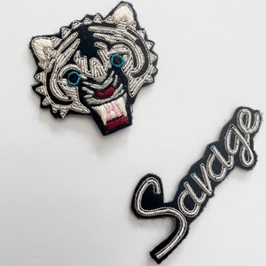 Peut inclure: Deux patchs thermocollants brodés. L'un représente une tête de tigre blanche avec des détails noirs et argentés et le mot "Savage" en noir avec des détails argentés.