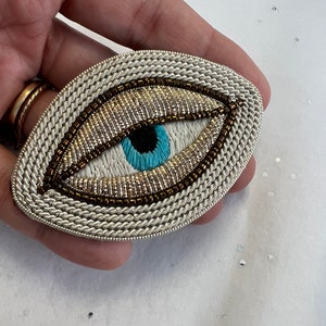 Op de afbeelding: Een witte ovale broche met een parel oogontwerp. Het oog is blauw met een zwarte pupil en een witte iris. De broche is omgeven door een zilveren koord.