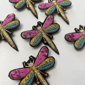 May include: Embroidered dragonfly patches in pink, gold, and teal. The dragonfly patches have black felt backing and are embellished with metallic thread. The wings are intricately detailed with a woven pattern. These patches can be used for clothing or craft projects.
