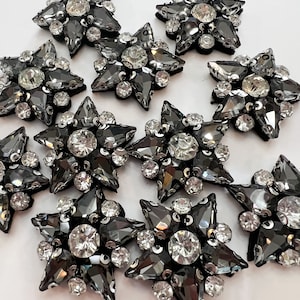 May include: A group of ten black star-shaped embellishments with clear rhinestones. Each star has a central rhinestone surrounded by smaller rhinestones.