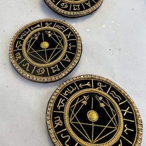 Könnte beinhalten: Drei runde Broschen aus schwarzem Samt mit goldener Stickerei. Jede Brosche zeigt ein goldenes geometrisches Design mit einem Kreis in der Mitte und einem Ring aus Tierkreiszeichen außen herum. Die Broschen sind von einem Ring aus klaren Strasssteinen umgeben.