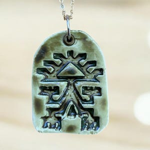 May include: A pendant necklace featuring a rectangular, olive-green ceramic pendant with a detailed, dark-green geometric design. The pendant hangs from a delicate silver chain. The design appears to be an ancient symbol.