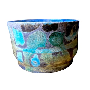 May include: A ceramic bowl with a textured, matte surface featuring an abstract pattern of mottled blue, turquoise, green, and dark brown patches. The interior of the bowl is a solid, vibrant blue, and the exterior base is a muted, earthy tan colour.