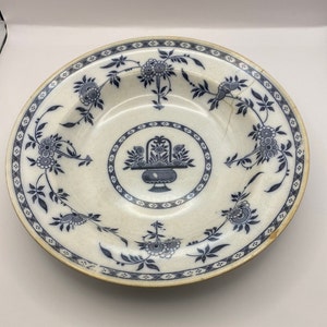 May include: A white ceramic plate with a blue floral design. The plate has a central design of a basket of flowers with a blue and white border.