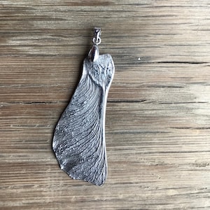 May include: A silver pendant shaped like a sycamore seed, with intricate veining details. It hangs from a small loop. The background is a weathered wooden surface.