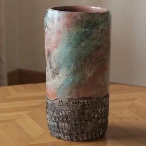 May include: A cylindrical ceramic vase with a textured, dark brown base and a colourful upper section. The upper portion features a blend of pink, green, and cream hues. The vase is displayed on a wooden table with a parquet pattern.