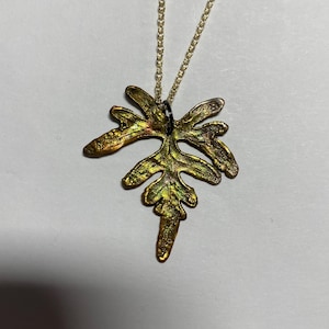 May include: A gold-colored leaf pendant necklace. The leaf has a textured surface with shades of gold, bronze, and green. The pendant is attached to a delicate gold chain. The necklace is displayed on a white background.