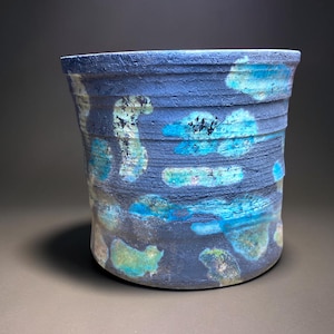Peut inclure: Un pot de fleurs cylindrique en céramique avec une finition mate bleu foncé texturée. La surface présente des stries horizontales et des taches irrégulières de glaçure turquoise vif, bleu clair et vert pâle créant un aspect organique.