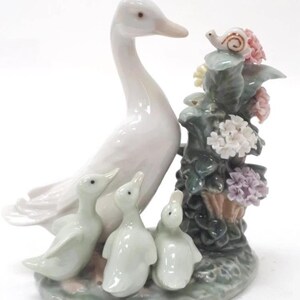 May include: A porcelain figurine depicting a white goose with three ducklings, alongside a floral arrangement with a snail. The goose is tall and elegant, with the ducklings clustered at its feet. The flowers are in shades of pink, white, and purple.