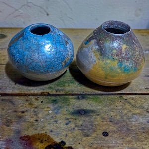 May include: Two small, round ceramic vases with narrow openings. The left vase features a bright blue crackle glaze finish. The right vase displays a mottled, multi-colored glaze with patches of yellow, green, brown, and metallic silver tones.