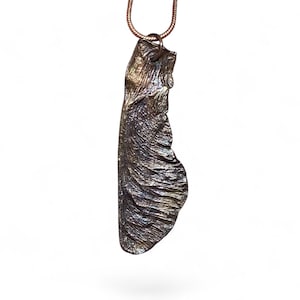 May include: A metallic pendant shaped like a maple seed pod, featuring a dark, textured, bronze-toned finish with subtle highlights. The pendant hangs from a thin, matching metallic snake chain, showcasing intricate vein-like details on its surface.