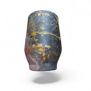 May include: A cylindrical ceramic cup featuring a dark charcoal gray base with mottled red and orange undertones. The surface is decorated with gold-colored splatters and a prominent horizontal gold-filled crack running across the upper section.