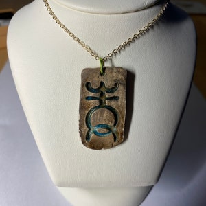 May include: A gold chain necklace with a ceramic pendant featuring a blue and white symbol. The pendant is rectangular and has a rough, textured surface.