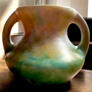 May include: A handcrafted ceramic vase with two loop handles. The vase features a blend of colors, including green, yellow, and orange hues. The vase is a decorative item, suitable for displaying flowers or as a standalone art piece.