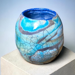 May include: A rounded ceramic vessel featuring a vibrant turquoise and blue glaze with a prominent dark crackle pattern across the exterior surface. The interior of the pot is finished in a solid deep blue, and the base is a natural tan clay colour.