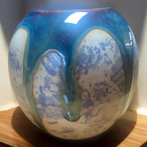 May include: A spherical ceramic vase with a wide opening. It has a blue glaze that drips down the sides, creating a unique pattern. The base colour is a light, speckled grey. The vase sits on a wooden surface.