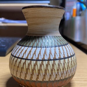 May include: A decorative ceramic vase with a textured surface. The vase features a brown rim and a patterned design in shades of brown, white, green, and orange. The vase is sitting on a wooden surface.