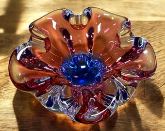 May include: A glass ashtray with a flower-like design in shades of pink, red, and blue. The ashtray has a deep center and a ruffled edge.