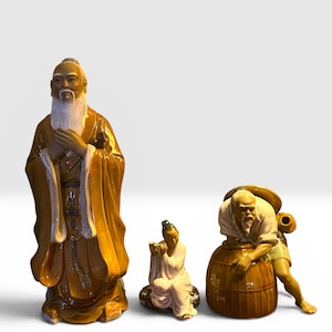 May include: Three ceramic figurines featuring a tall standing figure in a mustard-yellow robe with a long white beard, a smaller seated figure in a light pink robe playing a flute, and a figure leaning over a brown barrel with a straw hat.