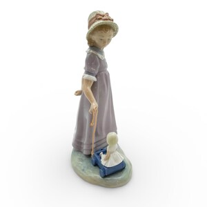 May include: A porcelain figurine of a girl in a lavender dress and a hat, pulling a blue wagon with a doll inside. The figurine is on a green and blue base. The girl is holding a rope attached to the wagon.