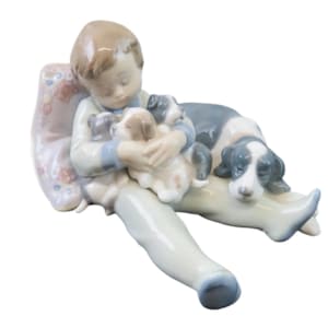 May include: A porcelain figurine of a child with puppies and a dog. The child wears a light blue and white outfit, seated on a pillow. The dog and puppies are white, grey, and brown. The figurine has a glossy finish, typical of porcelain.