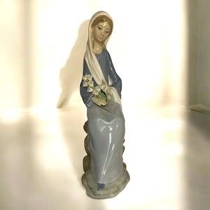 May include: A porcelain figurine of a woman in a blue dress and white shawl, sitting on a rock and holding a bouquet of white flowers.