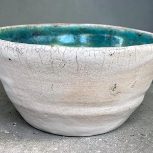 May include: A ceramic bowl with a rounded, slightly irregular shape. The exterior features a white glaze with a prominent dark crackle pattern throughout. The interior of the bowl is finished with a vibrant, glossy teal glaze that contrasts with the white.