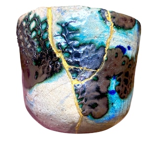 May include: A ceramic cup featuring a crackled, textured surface with a blend of turquoise, blue, and dark brown glazes. The vessel is repaired with prominent gold-colored lines that trace the fractures across its exterior body.