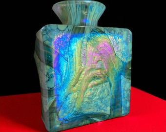 May include: A square-shaped, iridescent glass vase with a flared top. The vase features a blend of blue, green, and purple hues, with a textured surface. The design includes swirling patterns and a unique, artistic aesthetic.