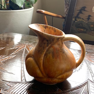 May include: A brown ceramic jug with a textured, organic design. The jug has a wide mouth and a handle. The glaze is a mottled brown and orange colour.