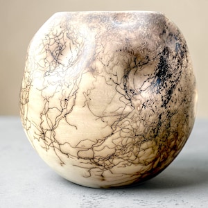 Könnte beinhalten: Eine kugelförmige Keramikvase mit einer glatten, cremeweißen, matten Oberfläche. Das Gefäß ist mit komplexen, dunkel schwarzen, verzweigten, haarähnlichen Mustern verziert, die sich um den abgerundeten Körper winden und einen Kontrast bilden.