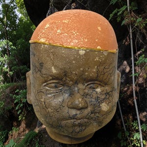 May include: A large, sculptural head figure featuring a smooth, terracotta-colored dome top separated by a thin gold band from the lower face. The face is a muted olive-brown color with a textured surface covered in intricate, dark, vein-like lines.