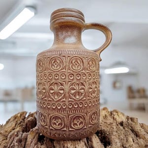 May include: A brown ceramic pitcher with a handle and a textured, patterned surface. The pitcher features a repeating floral design and is displayed on a piece of wood. The vintage-style jug is suitable for decorative use.