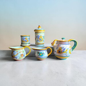 May include: A collection of five ceramic pieces featuring hand-painted designs in yellow, blue, green, and orange. The set includes a lidded canister, a teapot, two small pitchers with handles, and a cylindrical cup, all decorated with floral motifs.