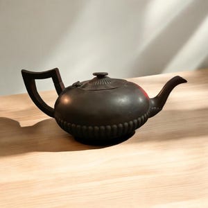 May include: A dark brown teapot with a curved handle and spout. The teapot has a round body with a ridged design around the base and on the lid. The handle is a dark wood color.