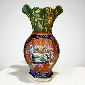 May include: A decorative ceramic vase with a ruffled rim. The vase features a green top with gold floral designs, a dark blue neck, and an orange body with painted floral and landscape scenes. The base is a dark blue, hexagonal shape.