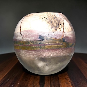 May include: A spherical ceramic vase featuring a multi-layered surface with horizontal bands of muted red, olive green, and dark charcoal. The upper section displays a cream-colored base with dark speckling and a prominent vertical crack.