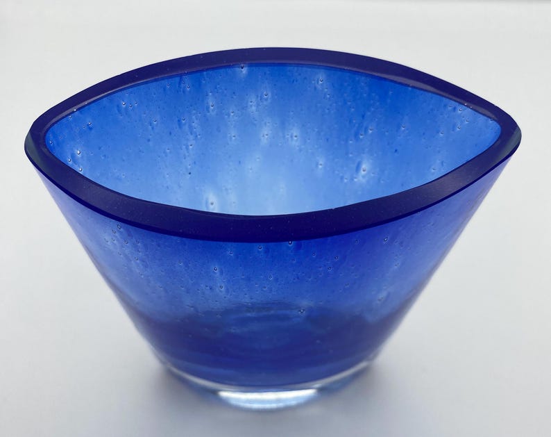 K&ouml;nnte beinhalten: Eine kleine, blaue Glassch&uuml;ssel mit leicht ovaler Form. Die Sch&uuml;ssel hat ein wei&szlig;es, wolkiges Aussehen mit einem dunkelblauen Rand.
