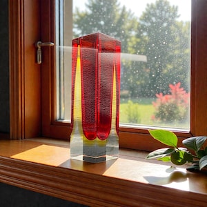 May include: A rectangular red and yellow glass vase with a clear base. The vase is on a wooden windowsill in front of a window. The vase has a geometric design and is lit by sunlight. The vase is approximately 20 cm tall.