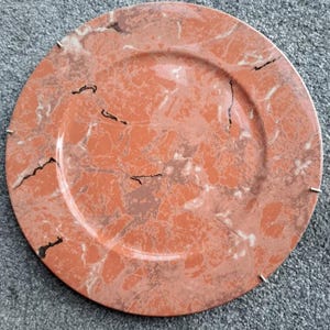 May include: A round, orange charger plate with a marbled design. The plate has a white rim and black veins throughout the design.