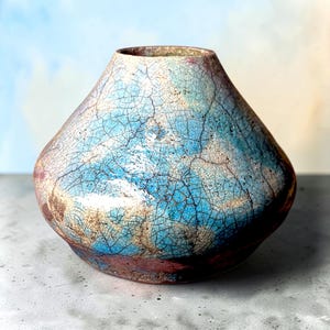May include: A wide, tapered ceramic vase featuring a prominent crackle glaze finish. The surface displays a vibrant blend of bright blue, white, and earthy brown tones, with dark, intricate web-like lines running across the entire exterior body.