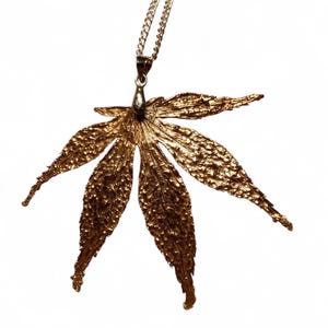 May include: A gold-toned pendant shaped like a seven-pointed leaf with a highly textured, rugged surface. The pendant is attached to a delicate metal chain by a small, smooth bail at the top, creating a detailed botanical jewellery piece.