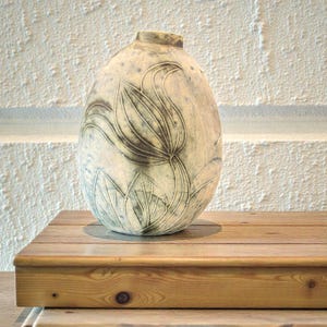 May include: A ceramic vase with a floral design. The vase is oval-shaped and has a light beige color with a darker floral pattern. The vase is sitting on a wooden surface. The background is a white textured wall.