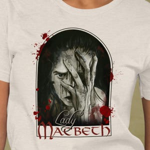 Peut inclure: Un t-shirt couleur crème arborant une illustration graphique d'une personne se couvrant le visage avec la main, entourée d'éclaboussures de peinture rouge. Le texte "Lady Macbeth" est imprimé en police noire stylisée en bas du motif.