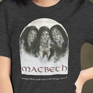 Pode incluir: Uma t-shirt cinzento-carvão escuro com uma impressão gráfica de três figuras encapuzadas com rostos pálidos e símbolos na testa. Abaixo das figuras, o texto diz MACBETH e something wicked this way comes numa fonte estilizada.