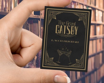 May include: A small, black book cover for "The Great Gatsby" by F. Scott Fitzgerald, held in a hand. The cover features gold art deco designs and two cocktail glasses. The background is a blurred bookshelf.
